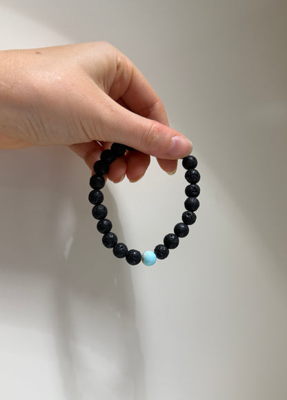 Hand holding a black beaded bracelet with a blue bead against a plain background