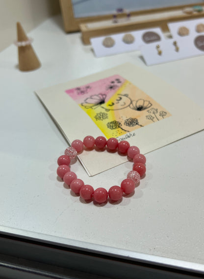 Pink beaded bracelet on a white surface with a card featuring a cartoon character in the background.
