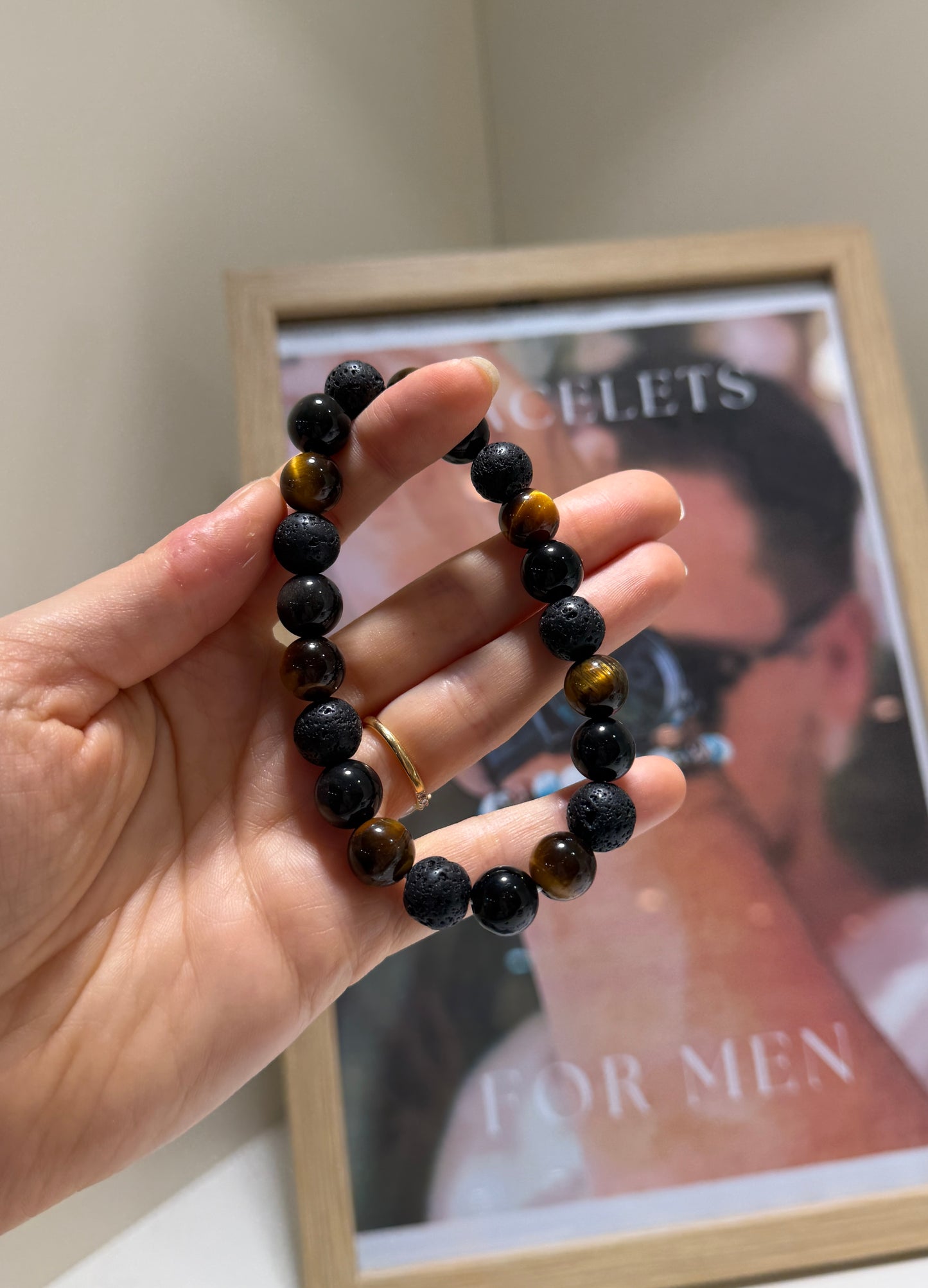Hand holding a black beaded bracelet with a blurred background featuring a magazine cover.
