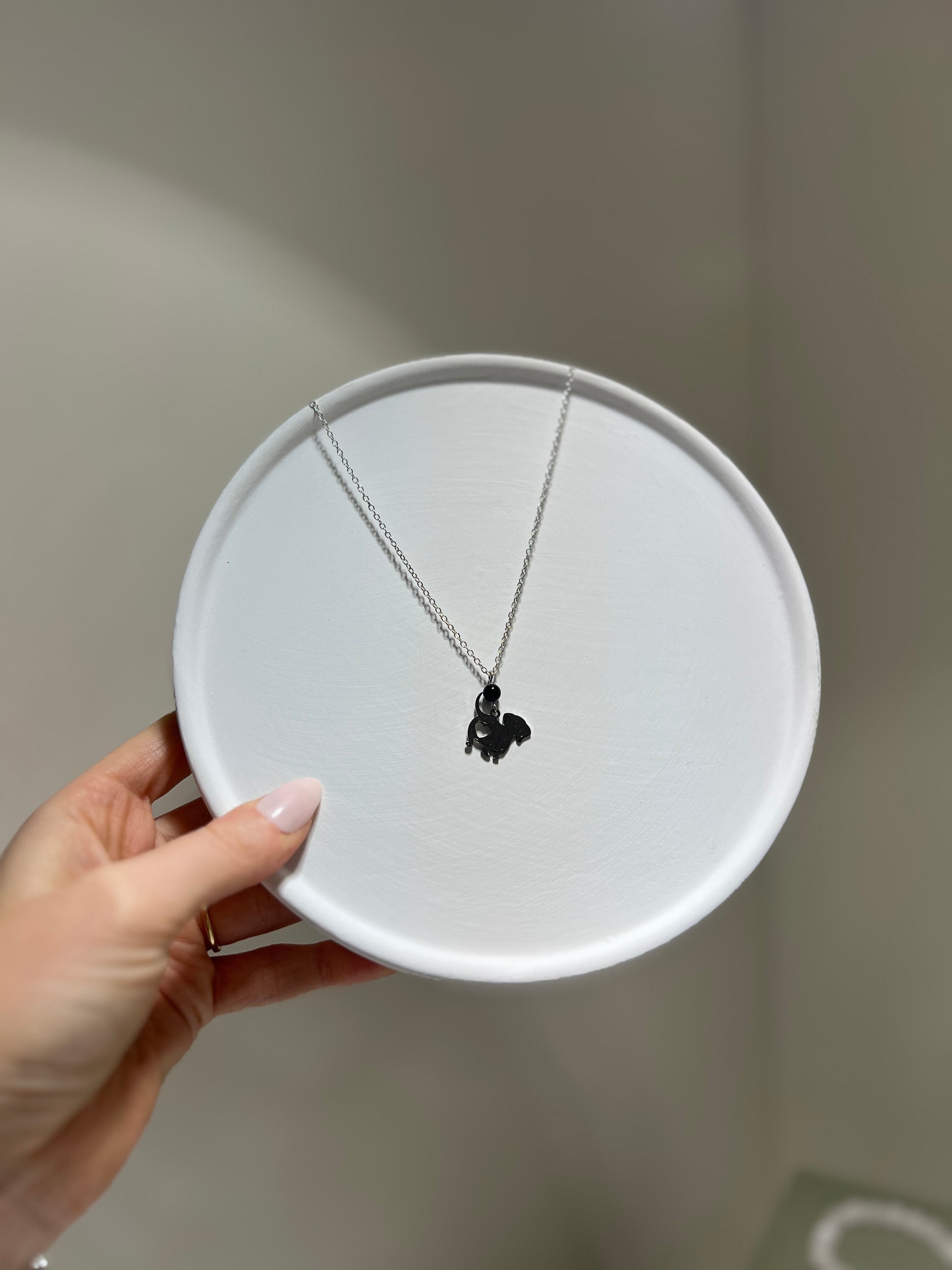 Necklace with a black pendant on a white round display stand held by a hand against a neutral background.