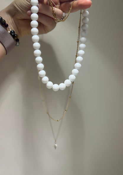 White beaded necklace and gold chain held by a hand against a neutral background