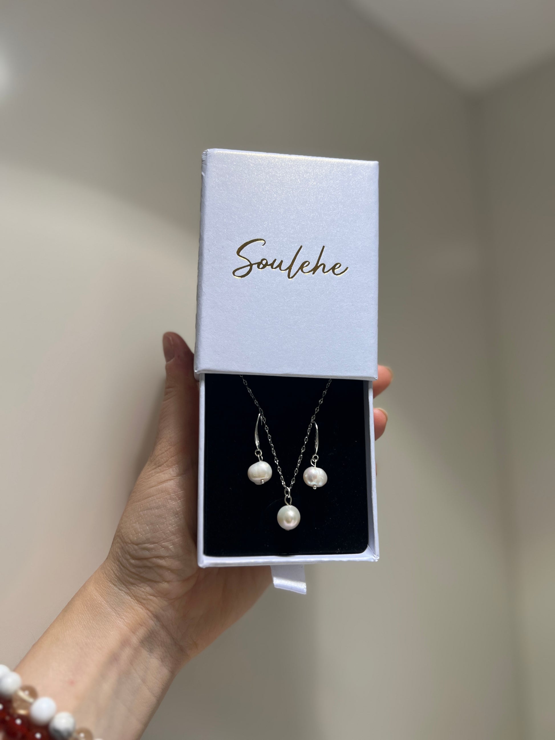 Pearl necklace and earrings set in a 'Soulehe' box held by a hand.