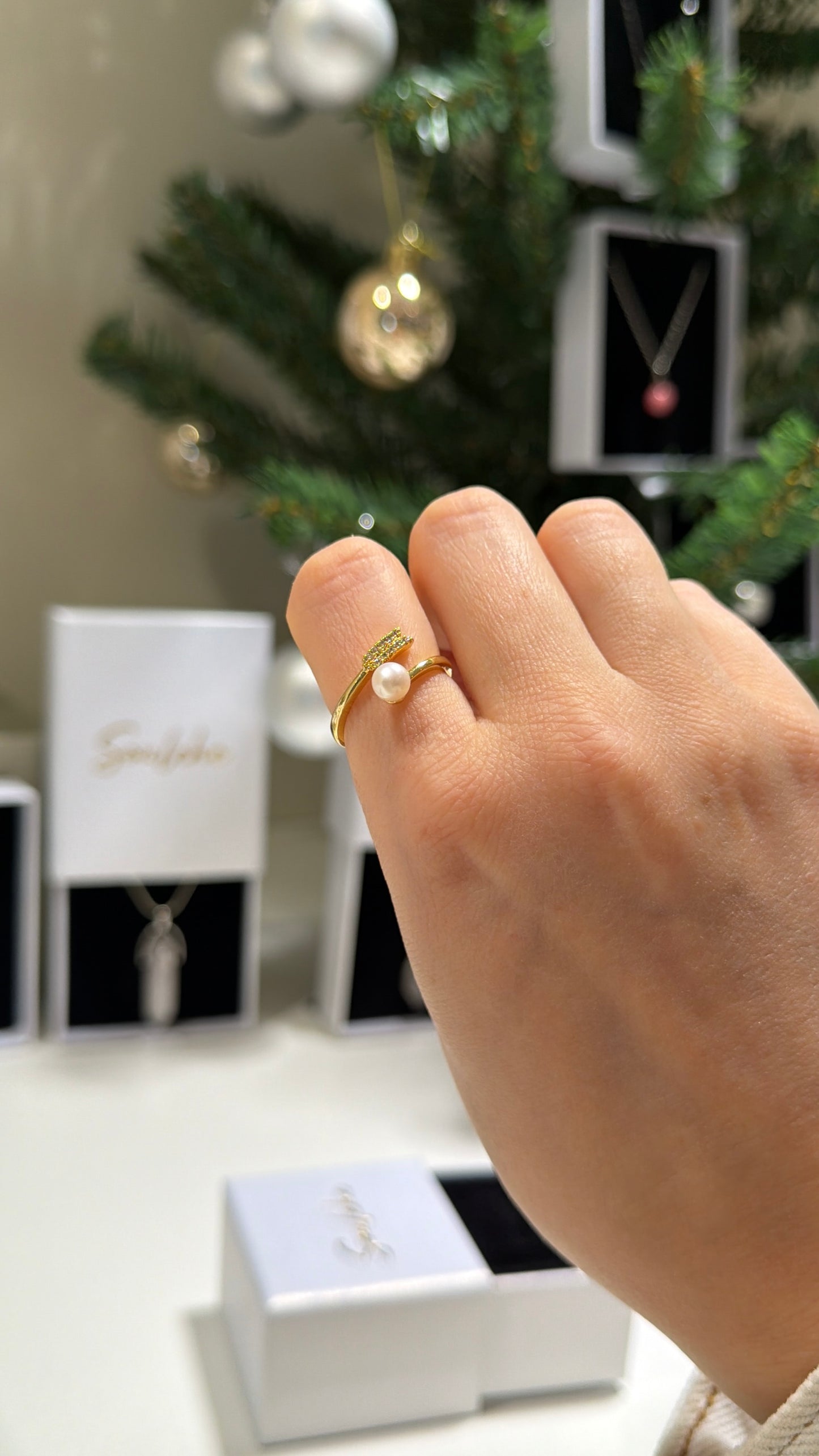 Hand wearing a gold ring with a pearl on a blurred Christmas tree background