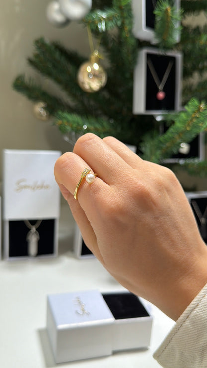 Hand wearing a gold ring with a pearl, with jewelry boxes and a Christmas tree in the background.